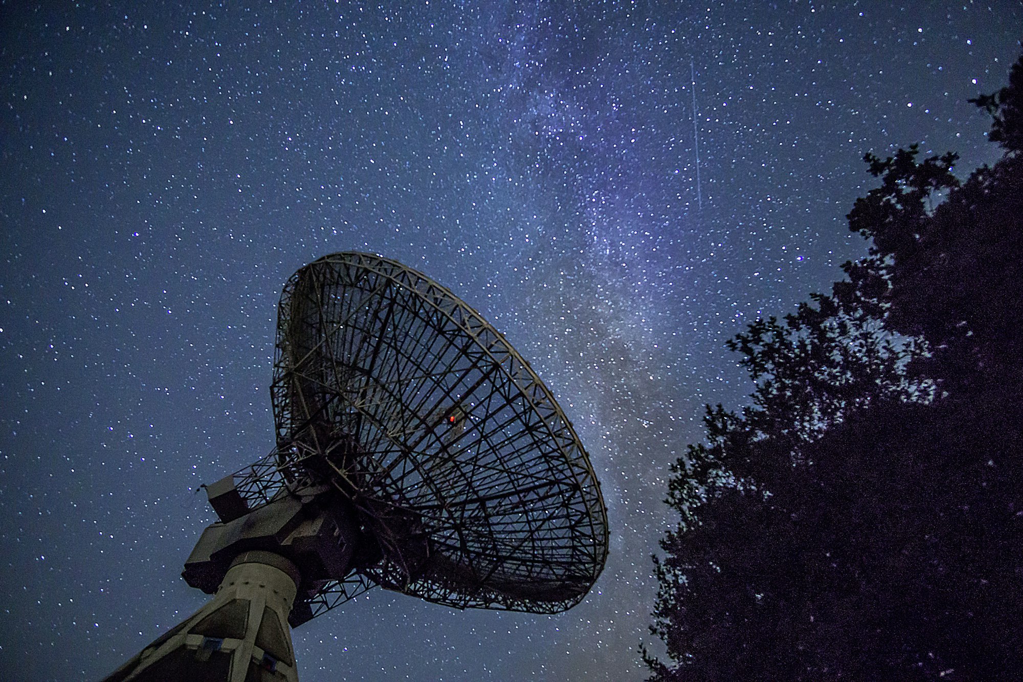 A radio telescope dish silhouetted against the Milky Way, one small red light on its rim